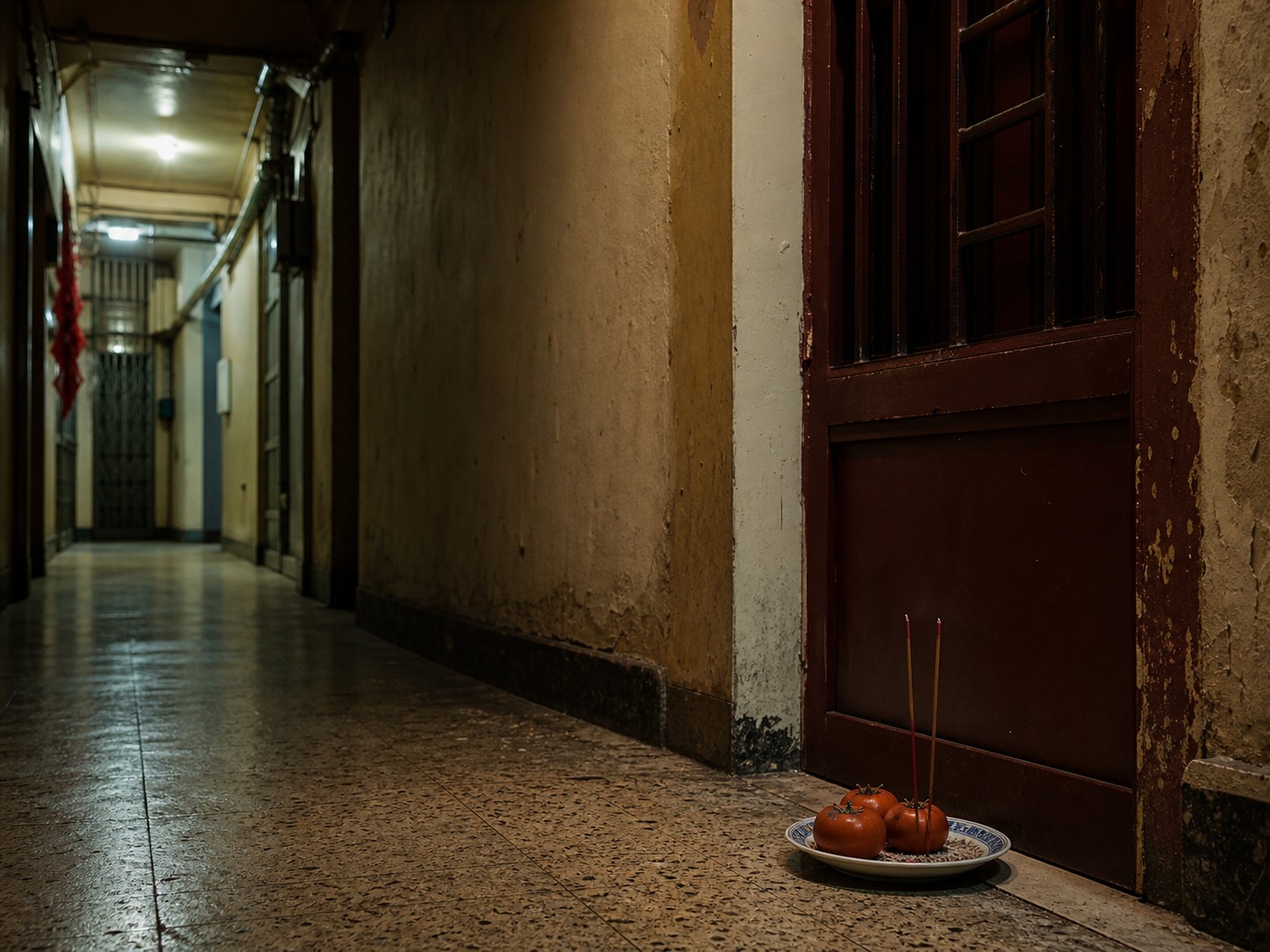 A dim apartment corridor with a small plate of oranges and incense sticks outside a closed door.