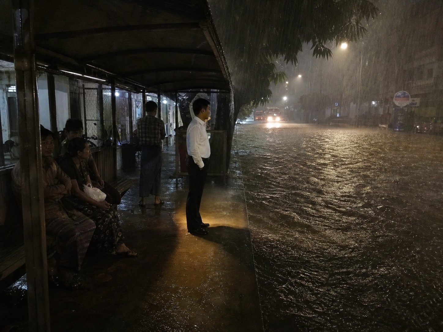 A rain-lashed Yangon street with a single man standing under a bus stop shelter, perfectly dry.