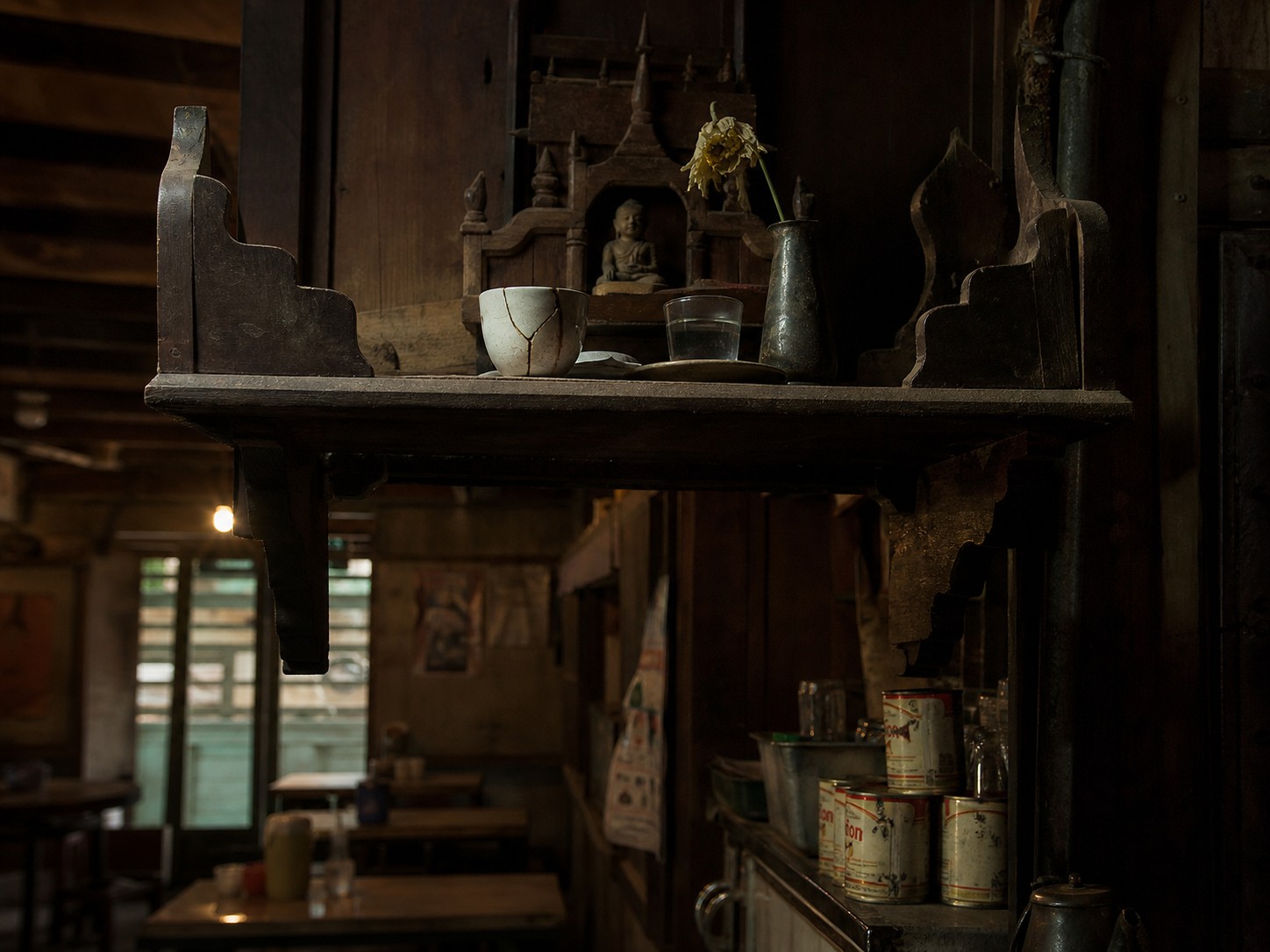 A dim tea shop interior with a high wooden shelf holding a cracked white cup and a single wilting flower.